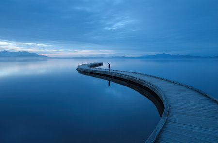 A solitary figure stands on a wooden pier extending into calm water, likely a lake or ocean. The scene features a monochromatic blue palette, with soft textures and reflections enhancing the serene atmosphere. The composition emphasizes the curve of the pier and the horizon line, suitable for various editorial and promotional purposes.の素材