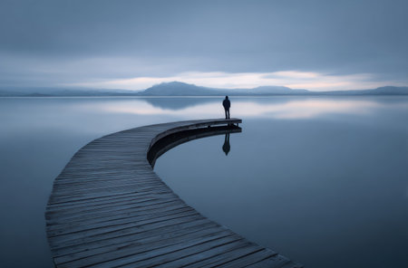 A person stands at the end of a wooden pier extending over a still body of water, possibly a lake. The scene is dominated by cool blue and gray tones, suggesting a cloudy day. The composition features a curved pier, leading the eye towards a distant mountain range. This image could be used for various commercial or editorial purposes.の素材