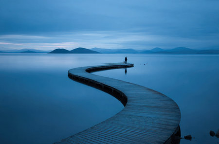 A wooden pier curves across tranquil water towards distant mountains under a dusky blue sky. A single person stands at the end of the pier, their silhouette suggesting contemplation. The composition features a symmetrical balance with soft lighting, suitable for various editorial and commercial applications.の素材