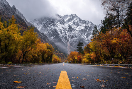 An asphalt road stretches into the distance, framed by vibrant autumn trees on either side. Towering, snow-covered mountains form the backdrop under an overcast sky. The image features a wide composition, capturing the natural beauty of the landscape. Potential uses include editorial illustrations or travel-related marketing.の素材