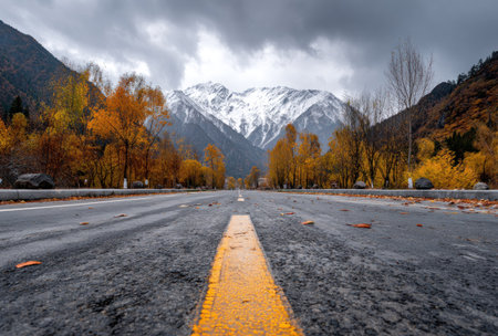 An asphalt road stretches towards a snow-capped mountain range, flanked by trees displaying autumn colors. The composition features a low-angle perspective, with the road markings in sharp focus. The lighting appears overcast, creating a muted color palette. Suitable for travel, nature, or environmental themes in various media.の素材