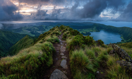 An aerial perspective shows a narrow path winding along a grassy hilltop, overlooking a serene lake and verdant landscape. The image features a cool color palette dominated by greens and blues, with dramatic cloudy skies overhead. This image could be suitable for travel, nature, or environmental content.の素材