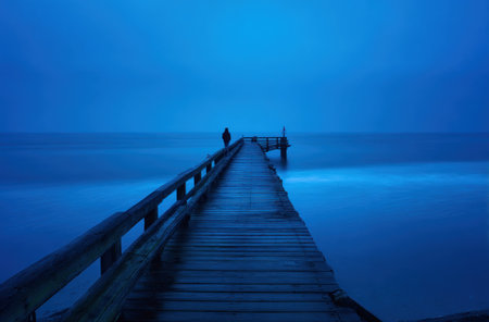 A person stands at the end of a wooden pier, silhouetted against the sea and sky. The image features shades of blue, with a long exposure creating a sense of movement. The composition suggests a calm and reflective atmosphere, suitable for various editorial and commercial applications.の素材