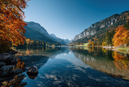 A serene landscape depicts a calm lake reflecting the surrounding mountains and trees. Warm autumn colors of orange and gold contrast with the deep blue sky. The composition features a natural outdoor setting, suitable for visual content related to nature and travel. The image is suitable for various commercial uses.の素材