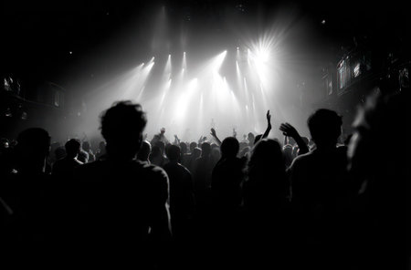 A crowd of people are silhouetted, enjoying a concert with bright stage lights overhead. The monochrome image presents high contrast, and a sense of energy and excitement. This visual may be suitable for promotional materials, editorial use, or imagery related to live entertainment. The setting appears to be indoors at night.の素材