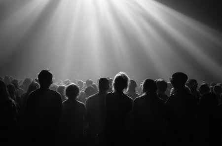 A crowd of people is silhouetted against a backdrop of powerful overhead light beams. The monochromatic image focuses on the collective, evoking a sense of unity and shared experience. The composition creates strong contrasts of light and shadow, potentially suitable for editorial content or design applications.の素材