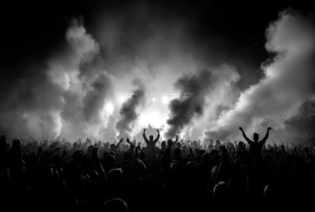A silhouetted crowd raises arms against a dark backdrop filled with billowing smoke. The monochromatic image showcases a dynamic composition with high contrast and overhead lighting. It evokes a sense of unity and excitement, potentially suitable for promotional materials or editorial content.の素材