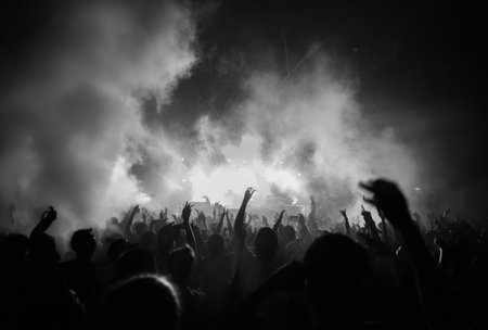 A grayscale image showcases a large crowd silhouetted against a backdrop of smoke or fog, with many arms raised in the air. The scene appears to be at night, possibly a concert or event, featuring dynamic composition and high contrast. The image could be used for various commercial or editorial purposes.の素材