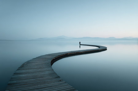 A curved wooden pier extends into tranquil water, under a pale blue sky. A solitary figure stands at the pier's end. The image uses a minimalist composition, featuring muted colors and a sense of calm. Suitable for various editorial and commercial applications such as travel or inspirational content.の素材