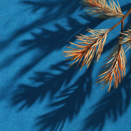 A close-up shot features a branch with golden-brown needles, casting dark shadows on a vivid blue backdrop. The overhead lighting creates a striking contrast between light and shadow. The composition highlights textures and shapes, suitable for various design and advertising purposes.の素材