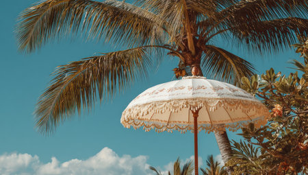 An ornate parasol stands beneath a large palm tree, bathed in sunlight against a backdrop of a clear blue sky. The image showcases soft colors and a slightly blurred focus. It may be suitable for illustrating themes related to leisure, vacation, or the tropics, with potential use in editorial or promotional contexts.の素材