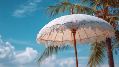 A cream-colored parasol, adorned with delicate lace, is featured against a backdrop of a clear, bright blue sky and palm tree. The image displays a soft focus with warm lighting that creates a relaxing atmosphere. This image is suitable for a range of commercial applications, including travel and lifestyle visuals.の素材