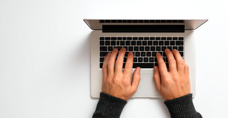 An overhead view displays hands actively typing on a laptop keyboard. The image showcases a modern laptop against a white backdrop, emphasizing the user's interaction. The lighting is bright and even, casting minimal shadows. Suitable for illustrating technology, work, or communication concepts.の素材