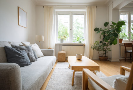 A well-lit living room interior showcases a comfortable sofa, chairs, and wooden furniture. Sunlight streams through a large window illuminating the space. The scene also contains houseplants and light-colored curtains that contribute to a peaceful atmosphere. Ideal for illustrating home decor or lifestyle concepts.の素材