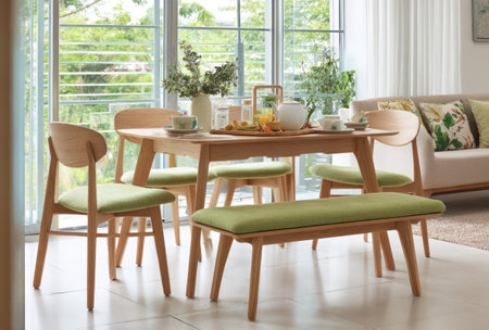 A well-lit interior scene presents a wooden dining table and chairs, complemented by a green bench. The composition showcases a domestic dining setting, enhanced by natural light filtering through large windows. This image could be used for various commercial and editorial applications to represent home interiors and lifestyle.の素材