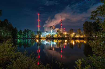An industrial facility is depicted at night with multiple smokestacks emitting vapors. The image showcases the complex's lights reflected in calm water, set against a dark sky. The scene is likely outdoors, featuring trees. This visual could be used for illustrations about industry or environmental topics.の素材