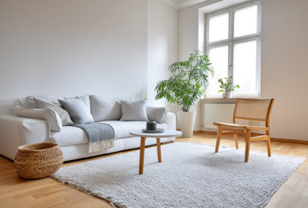 A bright living room features a white sofa, wooden chair, and small round table, complemented by a large window. A woven basket and potted plants add texture and natural elements. The interior uses neutral tones and natural light, suitable for lifestyle and home decor projects.の素材