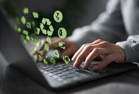 A person is using a laptop computer, with green icons of recycling symbols and environmental elements floating above the keyboard. The image features a close-up view with soft focus and a blurred background. The color palette primarily consists of green and gray tones, suggestive of concepts like sustainability or technology. This could be useful for eco-conscious or technology-related content.の素材