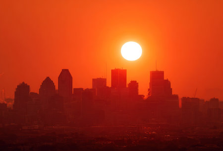 A cityscape is silhouetted against a brilliant orange sky, dominated by a large sun. Buildings create dark shapes, contrasting the bright backdrop. The overall image presents a warm, fiery aesthetic with soft lighting. This could be used for various commercial or editorial purposes.の素材
