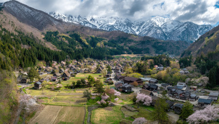 An aerial perspective captures a picturesque village situated in a valley, framed by mountains and forests. The image displays a vibrant mix of colors, featuring green vegetation, brown structures, and white mountain peaks. The composition suggests a serene setting, suitable for illustrating travel, nature, or community themes.の素材