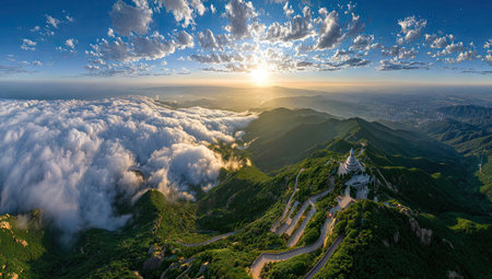 An aerial perspective showcases mountains partially obscured by clouds, with a bright sun radiating from the horizon. The landscape features shades of green and brown, with a winding road suggesting access. Suitable for use in a variety of commercial or editorial applications such as travel and nature content.の素材