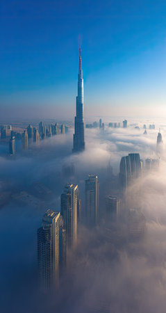 An aerial perspective showcases towering skyscrapers emerging from a sea of clouds. The composition emphasizes the contrast between the structures and the atmospheric conditions. The image features a clear blue sky, soft lighting, and potential applications in architectural, travel, or commercial projects. The scene evokes a sense of grandeur and scale.の素材
