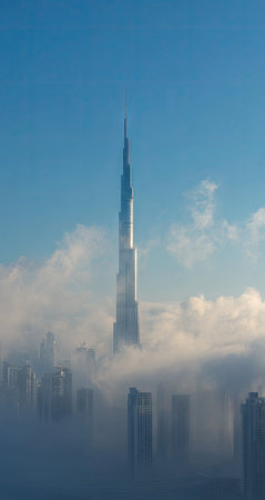 A tall skyscraper pierces through a layer of clouds, showcasing a modern architectural design. The image displays a clear blue sky, contrasting with the soft, white clouds. It presents a vertical composition with a sense of height and offers potential for commercial and editorial uses. This could be used for promoting business.の素材