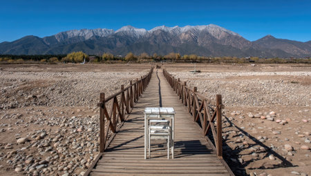 A wooden bridge leads toward distant mountains under a clear blue sky. A solitary chair sits on the bridge, the scene bathed in sunlight. The composition features a symmetrical perspective and natural colors. Suitable for various projects, it can be used to illustrate concepts of travel or isolation.の素材