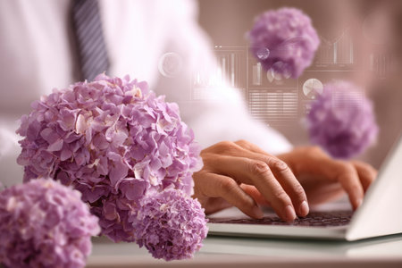 This image features a person working on a laptop, with focus on hands interacting with the keyboard. Soft, blurred floral elements in shades of purple are positioned near the hands. The composition shows elements of technology, nature, and the workplace. Suitable for various design projects and digital content.の素材