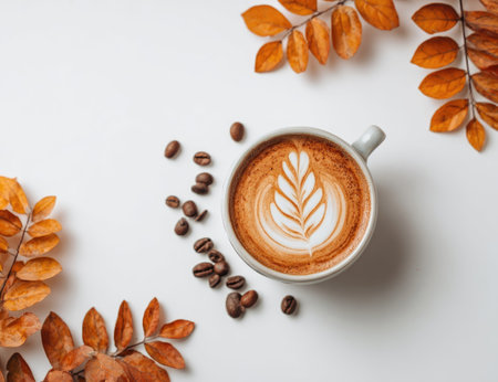 A top-down view showcases a cup of coffee with intricate latte art, accompanied by scattered coffee beans and vibrant orange autumn leaves. The image presents a clean composition, bathed in natural light, evoking warmth and a sense of seasonal change. Suitable for illustrating themes of relaxation and culinary enjoyment.の素材