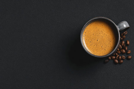 An overhead view displays a mug filled with coffee next to a scattering of roasted coffee beans. The drink is a light brown color, and the background is a solid black. This photo could be used for various commercial projects related to beverages or food products, enhancing marketing materials.の素材