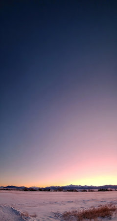 The image depicts a serene winter landscape under a colorful sky during sunset. The composition features a snow-covered foreground, with distant mountains silhouetted against a gradient of colors. The style is naturalistic with soft lighting. Suitable for environmental or travel-related publications.の素材