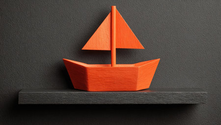 An orange paper boat sits on a dark gray shelf against a matching background. The image showcases simple geometry with bold color contrast and even lighting. The paper craft suggests a handmade quality, suitable for illustrative concepts related to travel, dreams, or creative design. Potential applications include website backgrounds and editorial content.の素材