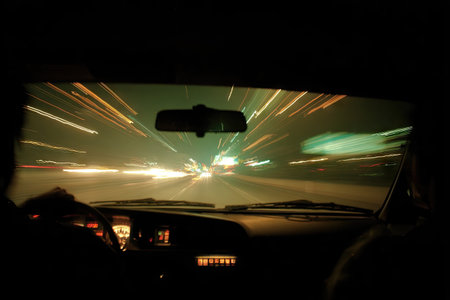 An interior car perspective captures a nighttime drive with blurred streaks of light. The composition features the dashboard and windshield framing the scene. Warm colors contrast with the darkness, suggesting speed and movement. Suitable for commercial applications, including transportation and travel concepts.の素材