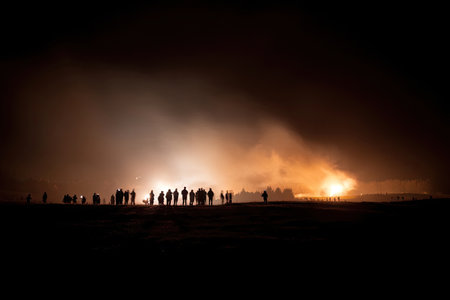 Numerous figures stand in silhouette, observing a bright, glowing event against a darkened night sky. The scene is illuminated by intense light, possibly a fire or explosion, creating a dramatic contrast. Suitable for editorial or commercial projects, the image conveys a sense of awe and mystery.の素材