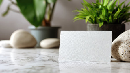 A clean white card stands upright, with blurred plants and stones in soft focus around it. The scene features natural elements of green and earth tones. The composition is likely for presentations, product showcases or marketing needs. The neutral setting allows for versatile applications.の素材