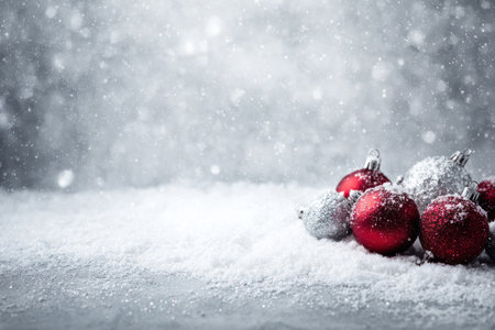Red and silver Christmas ornaments rest on a snowy surface in this wintry scene. The composition features a shallow depth of field, with soft focus on the falling snow. This image could be used for holiday-themed advertising, greeting cards, or editorial content, evoking feelings of festivity and celebration.の素材