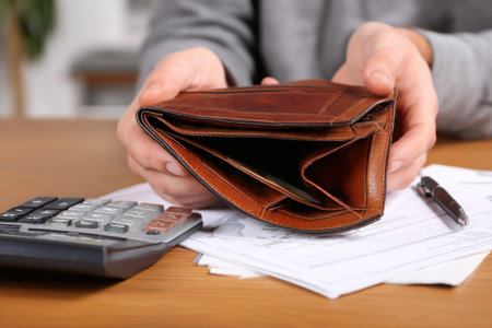 An empty brown wallet is held open, revealing its emptiness. A calculator and documents are visible on the wooden table, suggesting financial issues. The lighting is natural, casting shadows and highlighting the texture of the wallet. Suitable for various editorial and commercial projects related to finances.の素材