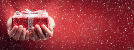 A pair of hands presents a gift box wrapped in red paper with a silver ribbon, against a blurred red background. White flakes create a falling snow effect adding a festive atmosphere. The image uses soft lighting to highlight the object, suitable for various promotional or seasonal visual content.の素材