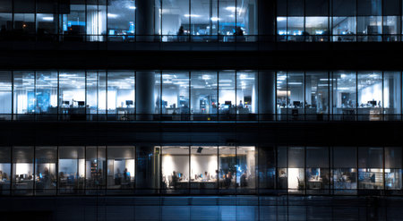 An exterior view captures a modern office building at night, with illuminated windows revealing interior workspaces. The composition showcases a grid of windows, suggesting multiple floors. The cool color palette emphasizes a nocturnal setting. Suitable for commercial or conceptual projects, the image evokes themes of work and business.の素材