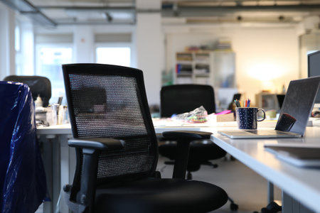 An office interior displays desks with laptops and chairs, suggesting a workspace environment. The composition features a neutral color palette with hints of blue. The arrangement offers a view of desks, chairs, and other office equipment, suitable for commercial projects or editorial purposes. The shot benefits from overhead lighting.の素材