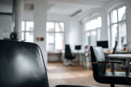 An interior shot features black chairs and multiple desks. The scene is bathed in bright daylight coming through large windows, giving a spacious and airy feel. The image conveys a modern and minimalist design. Suitable for articles or websites about business and office design.の素材