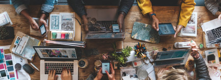An overhead shot depicts several people interacting with laptops, tablets, and smartphones on a wooden table. Warm lighting highlights the scene, suggesting an indoor environment. Various objects surround the devices, implying collaborative work or creative processes. The image could be used in business, technology, or editorial contexts.の素材
