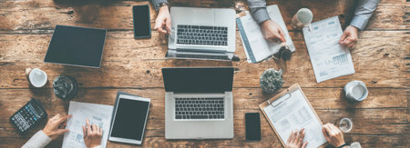 An overhead shot depicts a collaborative meeting with multiple laptops, tablets, and paperwork arranged on a wooden table. The warm tones of the wood contrast with the sleek screens. Papers, hands and devices suggest a business or creative setting, suitable for commercial projects.の素材