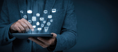 A person holds a tablet device with glowing digital icons hovering above. The image displays a professional setting with blue tones and soft lighting. It is suitable for conceptual illustrations related to technology, communication, and digital applications. The image can be used for commercial and editorial purposes.の素材