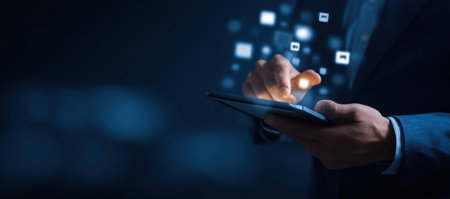 A person uses a tablet with a digital data overlay, set against a dark blue background. The image showcases glowing square icons floating above the screen. Ideal for illustrative uses in technology, business, or communication scenarios, the shot uses a shallow depth of field, with soft lighting and focus on the hands.の素材