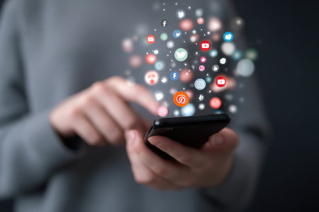 A person holds a smartphone, interacting with it. Floating above the device are various digital icons representing communication and social media platforms. The background is blurred, and the lighting is soft. This image is suitable for illustrating modern communication, technology, and related commercial topics.の素材