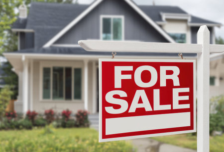 A "For Sale" sign dominates the foreground, framed against a blurred residential property. The sign is red and white, with bold text indicating availability. The background shows a house with a gray roof and windows, set within a well-maintained lawn. The image may be suitable for advertising, real estate, or housing-related content.の素材