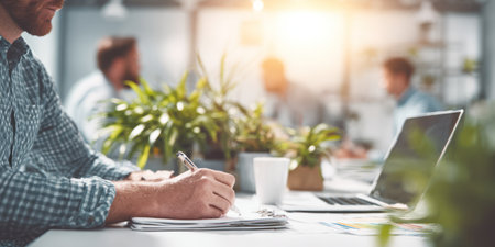 A person is writing on paper, near a laptop, in a bright office environment. The scene showcases a modern workplace with green plants and soft, natural lighting. Other individuals are blurred in the background, suggesting a collaborative atmosphere. Suitable for use in various commercial contexts, illustrating concepts of productivity and teamwork.の素材