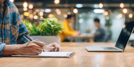A person is writing on paper with a pen at a wooden table, next to a laptop. The scene is indoors, likely a workspace or cafe, with blurred figures and greenery in the background. Overhead lighting creates soft illumination. Suitable for various commercial and editorial applications.の素材